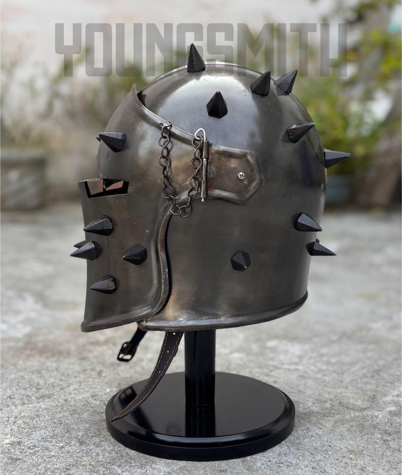 May include: A dark metal helmet with black spikes and a chain detail. The helmet is displayed on a black stand. The word "YOUNGSMITH" is visible in the background.