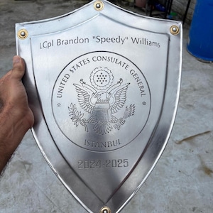Könnte beinhalten: Ein silberner Schild mit goldenen Nieten. Der Schild ist mit dem Text "LCpl Brandon Speedy Williams", "UNITED STATES CONSULATE GENERAL", "ISTANBUL" und den Jahren "2024-2025" graviert. Ein Adleremblem befindet sich in der Mitte.
