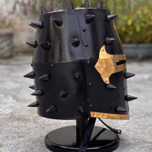 May include: A black metal helmet with numerous conical spikes. The helmet features a gold-colored decorative plate on the side and is mounted on a black stand. The helmet's surface has a glossy finish.