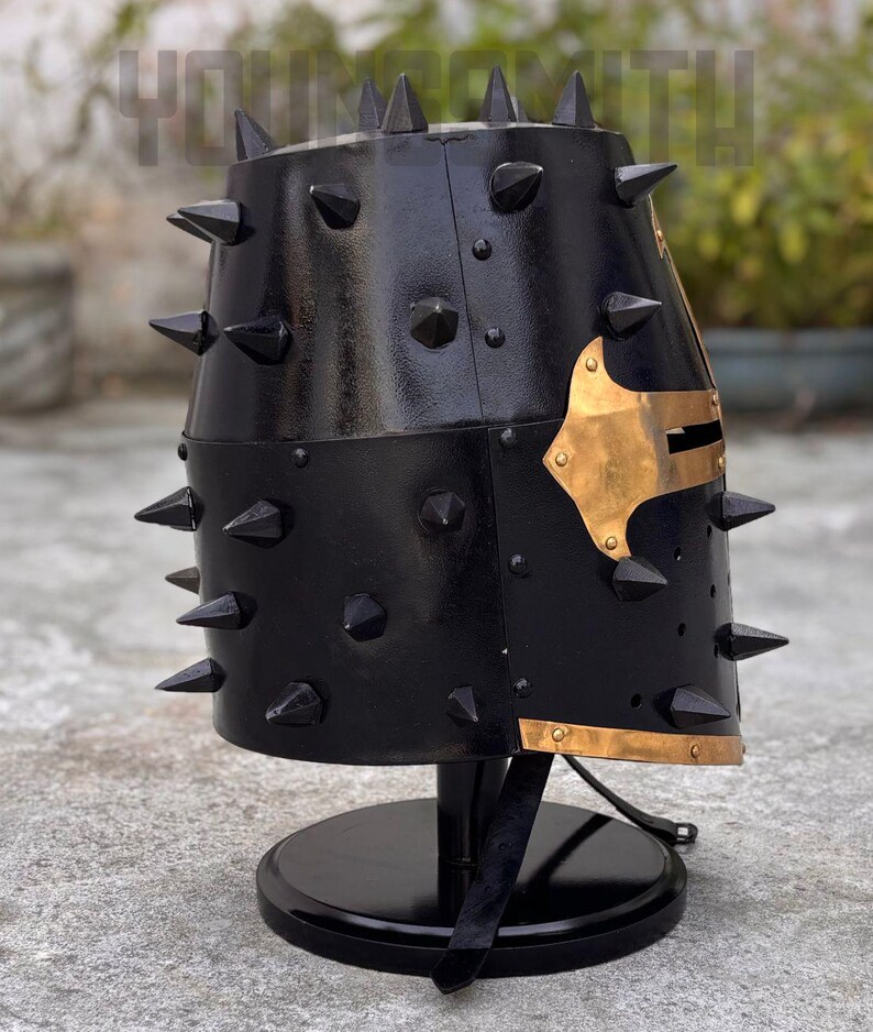 May include: A black metal helmet with numerous conical spikes. The helmet features a gold-colored decorative plate on the side and is mounted on a black stand. The helmet's surface has a glossy finish.