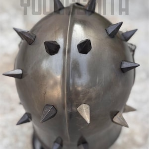 May include: A metallic, spiked helmet with the text "YOUNGSMITH" across the top. The helmet is a dark, muted color, with numerous dark, triangular spikes evenly spaced around its surface. The spikes are a darker shade than the helmet itself.