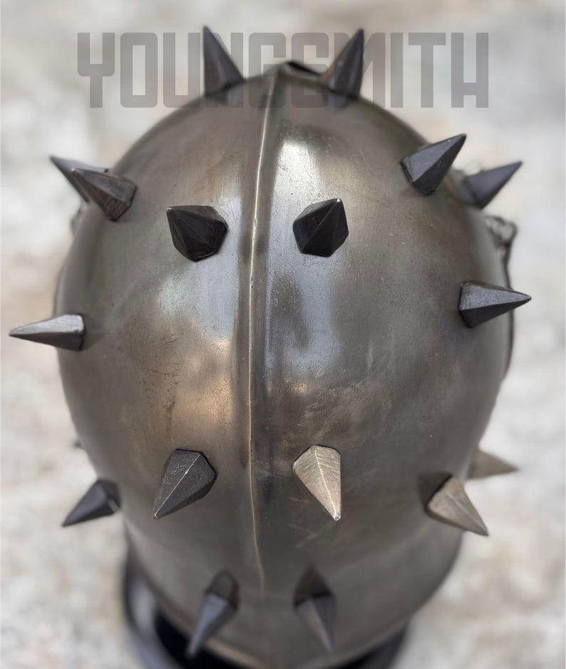 May include: A metallic, spiked helmet with the text "YOUNGSMITH" across the top. The helmet is a dark, muted color, with numerous dark, triangular spikes evenly spaced around its surface. The spikes are a darker shade than the helmet itself.