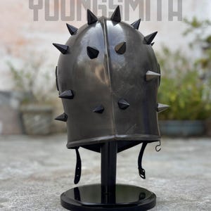 May include: A dark gray metal helmet with black spikes, displayed on a black stand. The helmet has a rounded top and a series of evenly spaced, pointed spikes. The text "YOUNGSMITH" is visible at the top.