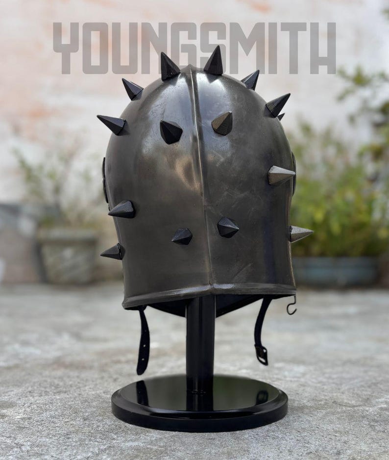 May include: A dark gray metal helmet with black spikes, displayed on a black stand. The helmet has a rounded top and a series of evenly spaced, pointed spikes. The text "YOUNGSMITH" is visible at the top.