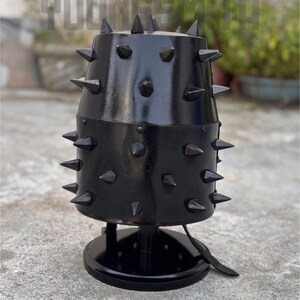 May include: A black metal lamp with a cylindrical shade, adorned with numerous pointed spikes. The lamp has a round base and a leather strap detail. The text "YOUNGSMITH" is visible in the background.
