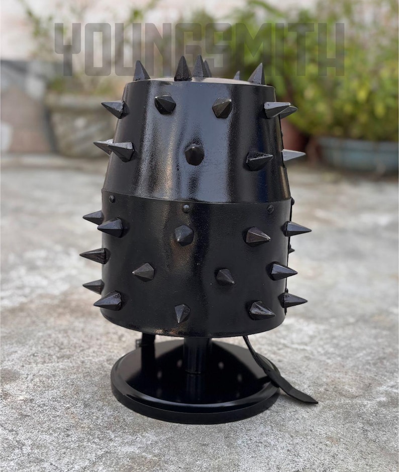 May include: A black metal lamp with a cylindrical shade, adorned with numerous pointed spikes. The lamp has a round base and a leather strap detail. The text "YOUNGSMITH" is visible in the background.