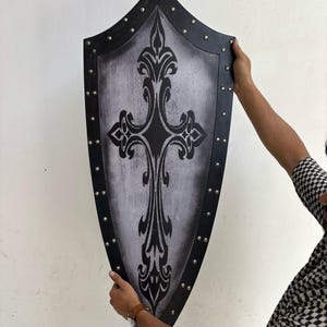 May include: A black and silver shield with a decorative cross design. The shield is made of wood and has a distressed finish. It is decorated with metal studs around the edge.