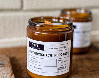 Może przedstawiać: Brązowa świeca w szklanym słoiku z etykietą "Butterscotch Pudding" z drewnianym knotem. Etykieta zawiera tekst "1971 candle co" i "soy wax candle". Świeca stoi na drewnianej powierzchni.