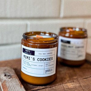 May include: A brown glass jar candle with a white label that reads "1971 candle co soy wax candle Mimi's Cookies" and "Used to all occasions" with a list of ingredients and "Handmade in Shelbyville, IL".