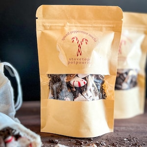May include: A brown paper bag with a clear window showing a mixture of chocolate, peppermint, and other ingredients. The bag has a round sticker with the text "spiced peppermint mocha stovetop potpourri" and a red and white candy cane design.