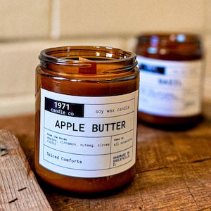 May include: A brown glass candle jar with a white label that reads "1971 candle co soy wax candle APPLE BUTTER Know the Notes apples, cinnamon, nutmeg, cloves Used to all occasions, rustic Spiced Comforts HANDMADE IN SHELBYVILLE IL".