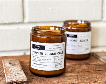 Może przedstawiać: Dwa bursztynowe szklane słoiki ze świecami z czarnymi etykietami. Świeca po lewej stronie jest oznaczona "1971 candle co. soy wax candle PUMPKIN CRUNCH CAKE" z tekstem "Pumpkin Everything" poniżej. Świeca po prawej stronie jest oznaczona "HOME AGACK".