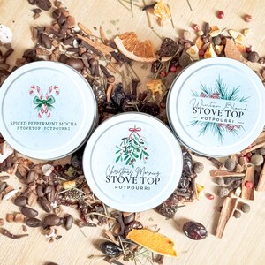 May include: Three round metal tins of stovetop potpourri with festive holiday designs. The tins are filled with a mixture of dried fruits, spices, and other natural ingredients. The labels on the tins read "Spiced Peppermint Mocha Stovetop Potpourri", "Christmas Morning Stovetop Potpourri", and "Winter Blend Stovetop Potpourri".