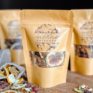 May include: A brown paper bag with a clear window displaying dried lemon slices, spices, and herbs. The bag is labeled "Lemon and Mint Leaf Stovetop Potpourri." Other bags and loose potpourri are visible on a wooden surface.