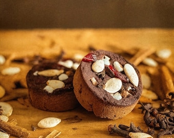 May include: Two round, dark brown baked goods topped with almonds and other ingredients sit on a wooden surface. Cinnamon sticks, cloves, and other spices are scattered around the baked goods, creating a warm, inviting scene.