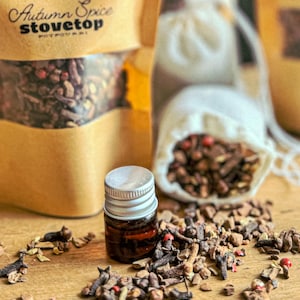 May include: A small brown glass bottle with a silver lid filled with essential oil. The bottle is sitting on a wooden surface with a variety of dried spices, including cloves, cinnamon sticks, and star anise. The spices are scattered around the bottle and a small white bag filled with spices is in the background. The bag has the text "Autumn Spice Stovetop Potpourri" printed on it.
