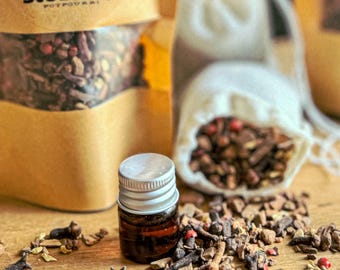 May include: A small brown glass bottle with a silver lid filled with essential oil. The bottle is sitting on a wooden surface with a variety of dried spices, including cloves, cinnamon sticks, and star anise. The spices are scattered around the bottle and a small white bag filled with spices is in the background. The bag has the text "Autumn Spice Stovetop Potpourri" printed on it.