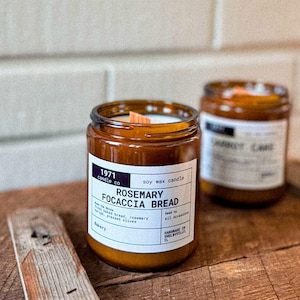 May include: Two amber glass candle jars with wooden wicks. The jar on the left has a white label with the text "1971 candle co soy wax candle Rosemary focaccia bread" and the text "Fresh baked bread, rosemary sprigs, pressed olives Bakery Handmade in Shelbyville, IL".