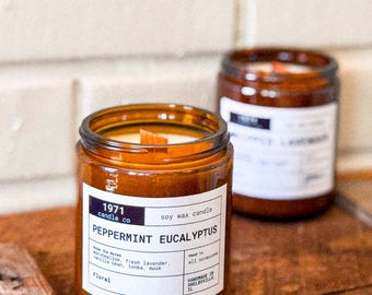 Może przedstawiać: Brązowa świeca w szklanym słoiku z białą etykietą, na której widnieje napis "1971 candle co soy wax candle Peppermint Eucalyptus". Etykieta zawiera również tekst "Know the Notes: Marshmallow, fresh lavender, vanilla bean, tonka, musk Floral" i "Handmade in Shelbyville, IL".