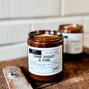 May include: A brown glass jar candle with a white label that reads "1971 candle co soy wax candle LEMON POSSET & PINE the notes: zesty lemon, sweet cream, sugar, pine needles, soft musk Bakery Handmade in SHELBYVILLE, IL".
