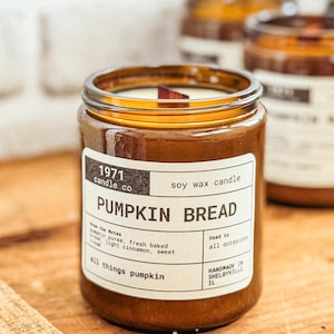 May include: A soy wax candle in a clear glass jar with a brown label. The label reads "PUMPKIN BREAD" and "all things pumpkin." The candle has a wooden wick and is handmade in Shelbyville, IL. The jar is sitting on a wooden surface.