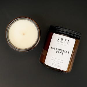 Könnte beinhalten: Eine Sojawachskerze in einem bernsteinfarbenen Glas mit schwarzem Deckel. Das weiße Etikett trägt die Aufschrift 1971 CANDLE CO. CHRISTMAS TREE, SOY WAX CANDLE, 8 OZ | 227 G, HAND POURED IN CENTRAL ILLINOIS. Eine zweite Kerze ist von oben zu sehen.