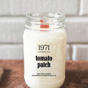 May include: A white glass jar candle with a black label that reads "1971 Candle Co. tomato patch Soy Wax Candle Handmade in Shelbyville, IL."