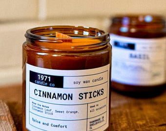 May include: A brown glass candle jar with a white label that reads "1971 candle co soy wax candle CINNAMON STICKS Know the Notes Cinnamon Leaf, Sweet Orange, Cinnamon Stick Spice and Comfort Used to all occasions HANDMADE IN SHELBYVILLE IL"