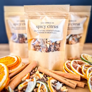 May include: A brown paper bag with a label that reads "1971 Candle Co. Spicy Citrus 100% All Natural Stovetop Potpourri | Handmade in Shelbyville, IL". The bag contains a mixture of dried citrus peels, cinnamon sticks, and other spices. The bag is sitting on a wooden table with dried orange and lime slices and cinnamon sticks scattered around it.