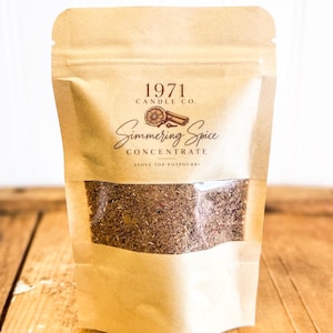 May include: A tan kraft paper pouch containing stove top potpourri. The label reads 1971 CANDLE CO. Simmering Spice CONCENTRATE STOVE TOP POTPOURRI with an illustration of a dried orange slice and cinnamon sticks. A clear window shows the contents.