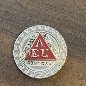 May include: A silver-coloured round pin with a red triangle in the centre. The triangle contains the letters "A" and "EU". The words "EDUCATE" and "DREAM" are on the outer edge of the pin. The word "CONTROL" is below the triangle.