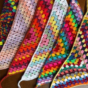 May include: A collection of colorful, handmade crochet bandanas. Each bandana features a granny square design in a variety of vibrant colors, including red, yellow, green, and purple. The bandanas have long ties for securing around the neck or head.