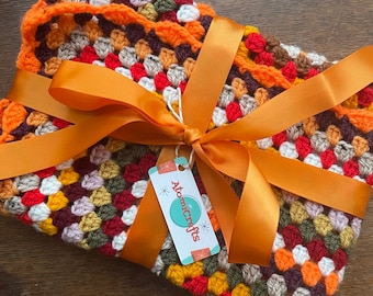 May include: A crocheted blanket in shades of orange, red, brown, white, and green is tied with an orange ribbon and a tag that reads "Atomicrafts".