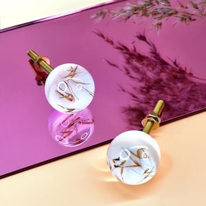 Peut inclure: Deux boutons de cabinet en résine blanche avec des tiges en métal doré. Chaque bouton présente un design unique de graminées et de fleurs séchées pressées.