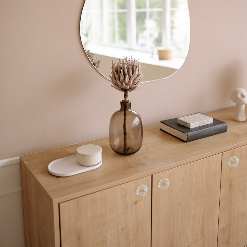 There is a view for a dresser in a beige room. Furniture is made from oak wood and has transoarent knobs with plants in it. On the cuboard are place a vase, a mirror and a book.