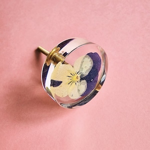 May include: A clear glass knob with a pressed purple and yellow flower inside. The knob has a brass screw on the back.
