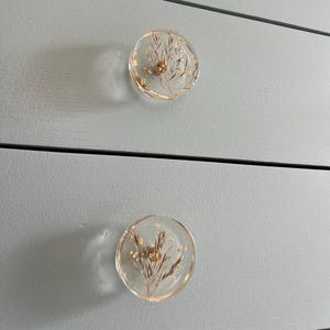 Könnte beinhalten: Zwei runde, transparente Schubladenknöpfe aus Harz mit getrockneten Blumen und goldenen Akzenten.