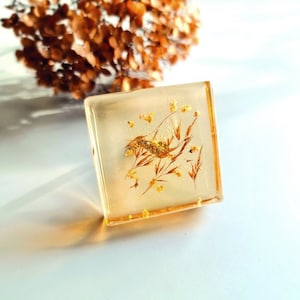 Può includere: Un fermacarte quadrato in resina trasparente con fiori secchi e glitter dorati all'interno.