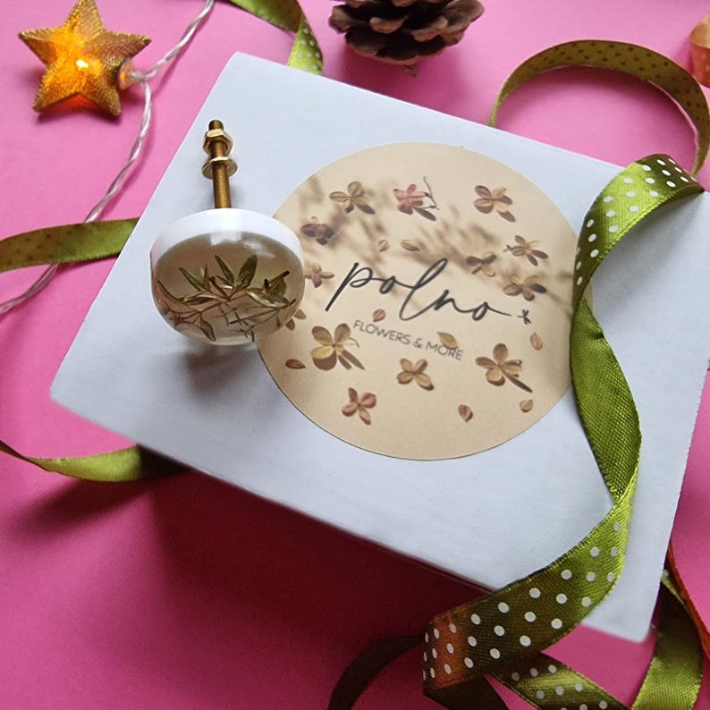 May include: A white ceramic knob with a gold metal stem and a pressed floral design. The knob is on a white box with a brown circle sticker that says "polno" and "flowers & more".