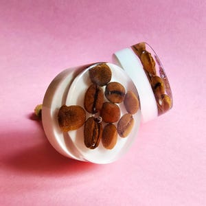 Könnte beinhalten: Zwei weiße runde Knöpfe mit klaren Harz-Oberteilen, gefüllt mit Kaffeebohnen. Die Knöpfe haben eine glatte, glänzende Oberfläche.
