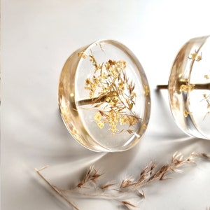 Transparent clear knob, round shape, with brass thread. In this drawer pull there are plants like: ears of grass and white lilac petals. Furniture handle is placed on a white table.