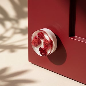 May include: A clear, round cabinet knob with embedded red raspberries. The knob is mounted on a deep red cabinet door. The raspberries are visible through the clear resin, creating a unique and decorative accent.