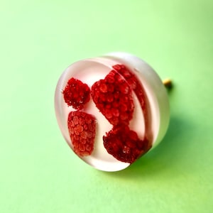 Può includere: Un anello di resina trasparente con tre lamponi rossi incorporati al suo interno. L'anello è su uno sfondo verde.