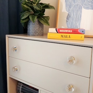 Light grey dresser standing in the bedroom. It has transparent clear drawer pulls with dry plants in it.