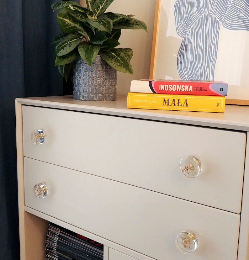 Light grey dresser standing in the bedroom. It has transparent clear drawer pulls with dry plants in it.