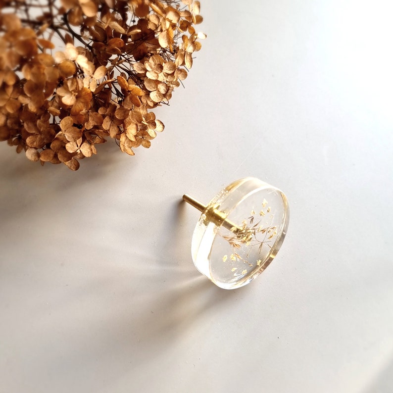 Transparent clear knob, round shape, with brass thread. In this drawer pull there are plants like; ears of grass and white lilac petals. Furniture handle is placed on a white table.
