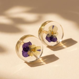 Op de afbeelding: Twee heldere, ronde oorbellen met een gouden rand, elk met een geperste, gedroogde bloem. De bloemen hebben paarse en gele bloemblaadjes. De oorbellen zijn geplaatst tegen een neutrale achtergrond met zachte schaduwen.