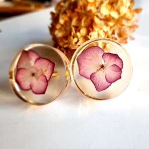 Puede incluir: Dos pendientes de resina transparente, cada uno con una flor rosa prensada en su interior. Las flores son de color rosa claro y tienen un aspecto delicado y en capas.