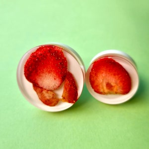 Puede incluir: Dos pendientes redondos de resina transparente con rodajas de fresa roja en su interior. Los pendientes están sobre un fondo verde.