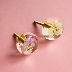 Op de afbeelding: Twee transparante glazen kastknoppen met een messing schroef. Elke knop heeft een geperste bloem erin, een met een roze bloem en de andere met een witte bloem.